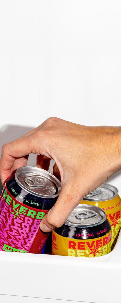 A hand reaching into a fridge grabbing a can of Reverb Splash.
